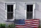  oil painting of a white clapboard house,two windows with flag draped along bottom sills, weeds along base