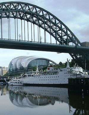 An iron bridge with a semi-circular upper structure, over the river Tyne. Beneath the bridge is a large white boat with several decks.