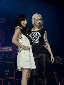 Two 24-year-old women are standing onstage with arms around each other. The left woman is shown in right profile, she has dark hair and a light-coloured dress with her right arm on her hip. The right woman is looking to her left, she has light hair and a dark T-shirt with a white design which includes a skull-and-crossbones and the words "Short Stack". She wears a large black cross on a dark chain which hangs over her T-shirt. Her left upper arm has tattoos. Both women wear dark red lipstick.