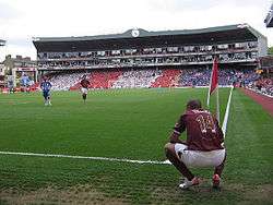 A coloured photograph of a man, crouching down on the right side. He is wearing a redcurrant shirt with white shorts; the shirt has 'Henry' and the letter 14 printed on the back. The landscape of the photograph is a filled section of a stadium.