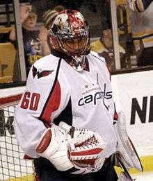 Hockey player in white Washington Capitals uniform with the number sixty on it, and goaltender's gear. He stands at ease in front of goal, hands by his side, and looks downward.