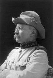 Profile of a white man, arms crossed, with drooping mustache and pince-nez glasses wearing a Hardee hat and a military jacket with the letters "U.S.V" on the upright collar.