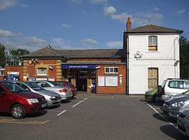 A brown-bricked building with a rectangular, dark blue sign reading "THEYDON BOIS STATION" in white letters all under a blue sky with white clouds