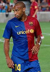 A dark-skinned man wearing a blue and red shirt and blue shorts with the number "14" displayed on the lower right thigh.