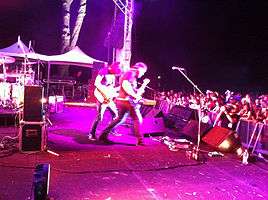 Side of stage shot of a band playing. Two members are shown in right profile both are playing guitars. Other stage equipment is visible around them. The audience is to the right and below stage level.