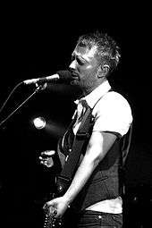 Black and white image of a man wearing a white dress shirt, a dark vest and jeans holding a guitar and standing behind a microphone stand. His eyes are closed, and the background is completely black except for a single light that shines from behind.