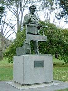Bronze statue of a standing man in military uniform and peaked cap atop a stone base with a plaque at the front. He is clasping the window frame of a jeep.