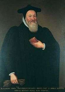 A man with full beard, wearing a black pointed cap and robes. He is holding a book in his left hand against his body, and a pair of gloves in his right hand by his side