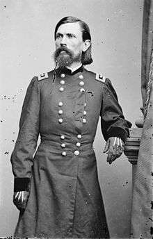A man in his late thirties with black hair, a mustache, and a long, black beard. He is wearing a long military coat and gloves and is facing right.