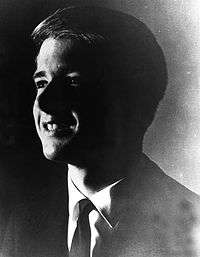 Shadowy profile of smiling young white man wearing a dark suit coat over a white shirt and dark tie.