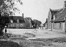 Large thatched cottage on right, man standing in front of pond on left, large thatched cottage in background with a small group of people and two horses standing nearby