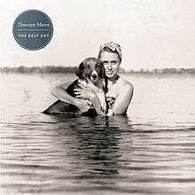 A black-and-white photograph of a woman in water holding a dog. In the upper-left corner, white text inside a teal circle reads "Thurston Moore" in standard case, followed by "The Best Day" in uppercase text.