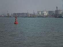 A red port-side buoy with an oil wharf and tankers in the background