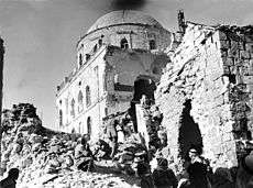 Black & white image showing gaping hole in side of synagogue with soldiers walking atop rubble outside