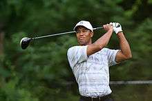 Tiger Woods, an African-American male in his early-30s, looks serious and wears a white hat and blue greyish polo shirt. On both his hands, he holds a golf club, after swinging it, and sports a white glove on his left hand.