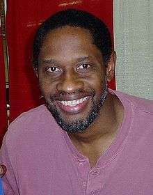 A dark-skinned man with black hear and a greying beard and moustache is wearing a violet shirt and smiling into the camera.