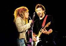 Turer and Clapton, on stage, sharing a microphone stand, singing.