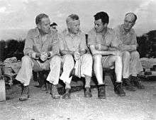 Four men in rumpled fatigues sit on the ground. Tibbets is wearing shorts.