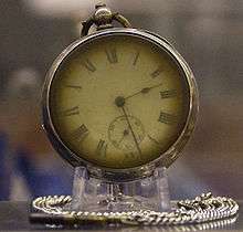 Photograph of a brass pocket watch on a stand, with a silver chain curled around the base. The watch's hands read 2:28.