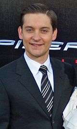 An image of a Caucasian man smiling. He has medium brown hair and is wearing a navy jacket over a white shirt and striped tie.
