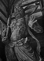  Agyō. Front view of a scary statue carrying a spear like object in his right hand and the palm of his left hand facing the viewer with fingers spread. Sculpted breastplate and necklace are visible.