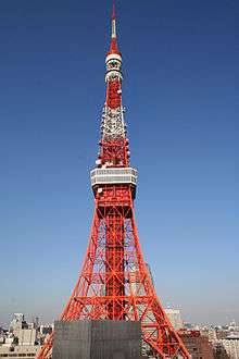 The orange and white lattice frame of Tokyo Tower rises up in front of a clear, blue sky.