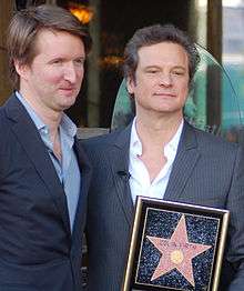 Two middle aged men stand side by side wearing suits and open-necked shirts. One is holding the plaque of a Hollywood star of fame