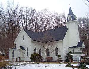 A white church with a black roof, pointed windows and a pointed steeple, with woods full of bare trees in the back. There are a few patches of snow on the ground in front.