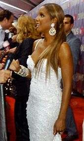 Photograph of a woman being interviewed by a reporter. She has long blond hair that is combed with a pony tail. The woman wears a long white dress, which has diamonds throughout. Accessories she wears are diamond bracelets, a ring and earrings.