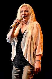 Colored photograph of Toni Willé standing onstage holding a mike. She has long blonde hair and is wearing a black top and off-white full sleeved top with grey pants.