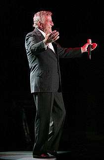 An older man holding a microphone in one hand, his arms held out, smiling and wearing a black suit with a white dress shirt.