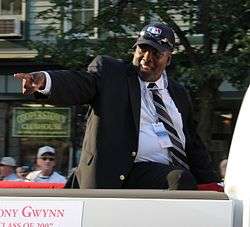 Profile view of a dark-skinned, heavyset man wearing a dark suit, white dress shirt, and a ballcap. He is in an elevated, sitting position in a car pointing to the left with his right arm.