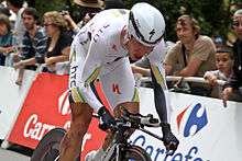 A road racing cyclist, wearing a predominantly white skinsuit with black, green, and gold trim, and an aerodynamic helmet. He is crouched low, but riding out of the saddle. Spectators watch from behind roadside barricades.