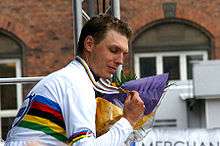 A man in his late twenties wearing a mostly white cycling jersey with rainbow stripes across the midsection and biceps. He has a medal hung around his neck, and clutches it with his right hand. In the other hand is a bouquet of flowers.