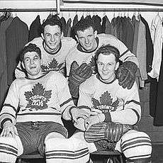 Four hockey players, two sitting and two standing behind them, smiling triumphantly.