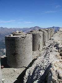 A row of round brick turrets can be seen in the foreground. A mountain range occupies the background.