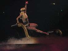 Image of a blond woman. Her right hip is resting on a black divan, with her right leg extended to the end of the divan. Her left hand is extended in the air, while her right hand is grabbing the top of the divan. She is wearing a flesh-colored body suit with tattoos and nipple tassles. She is sporting a black blindfold and singing into a wireless microphone. The divan is surrounded by smoke.