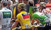 Four road racing cyclists viewed from behind, wearing special jerseys that are, from left to right, prominently white, yellow, green, and red polka-dotted.