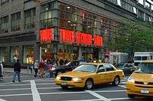 A street view of a store front prominently features a yellow taxi in front of the store. The walls contain clear windows with the phrase "Tower Records - Video" in bright orange lights.