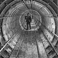 Etching of a circular tunnel made of bolted segments. A walkway of boards runs down the centre of the tunnel and a man in workman's clothes with a bag over his shoulders stands in the middle distance