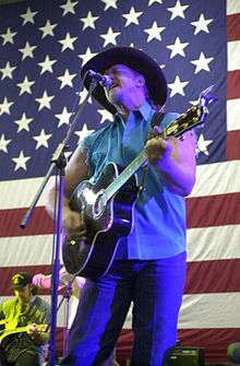 A man with his eyes closed, singing into a microphone, and playing an acoustic guitar. He is wearing a cowboy hat with blue jeans and a sleeveless blue dress shirt. A large American flag appears in the background, as do two band members.