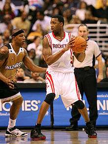 A basketball player, wearing a white jersey inscribed with the word "ROCKETS" across the front, holds a basketball away from another basketball player guarding him. A referee stands in the background.
