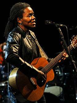 A woman with dreadlocks standing behind a microphone stand. She is wearing a leather jacket and playing a guitar.