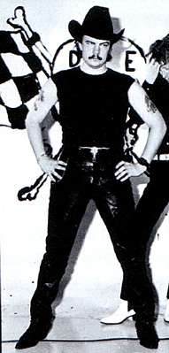 A 23-year old man stands akimbo, facing forward. He wears a black cowboy hat, sleeveless t-shirt, leather pants, a band on his left wrist and pointed shoes. He has a moustache and has tattoos on his upper arms. He is in front of a sign/art work which is mostly obscured but with a chequered flag, bones and the letters D and E visible. To his left and behind him is another man partly cut off at the edge of the image.