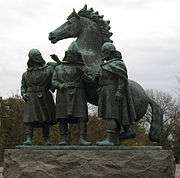 Photograph of a copper statue (coated with green patina) of three men casually discussing with a horse in the background.