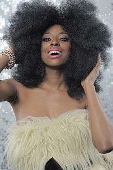 A dark-skinned woman wearing heavy make-up is smiling towards the camera. She is touching her large, black hair, and is wearing a furry, whte top.