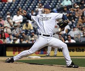 A man in a white baseball uniform and navy blue cap stands on a dirt mound throwing a pitch with his right hand. He is wearing a black belt, black shoes, and a black baseball glove, and his uniform reads "Padres" in navy blue script across the chest, outlined in tan. His cap has two interlocked letters: "S" and "D".