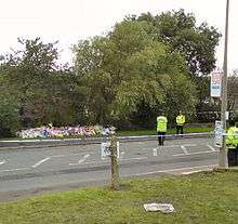 Three policeman stand near a large number of bouquets beside a road