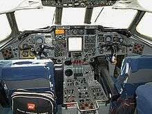 The flight deck of a jet airliner with many instruments and controls. Two pilots' seats have small bags hanging on them, one showing the words "British European Airways".