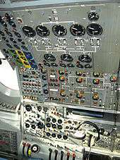 An airliner flight deck control panel with many instruments and switches. A pair of headphones hang from the panel.